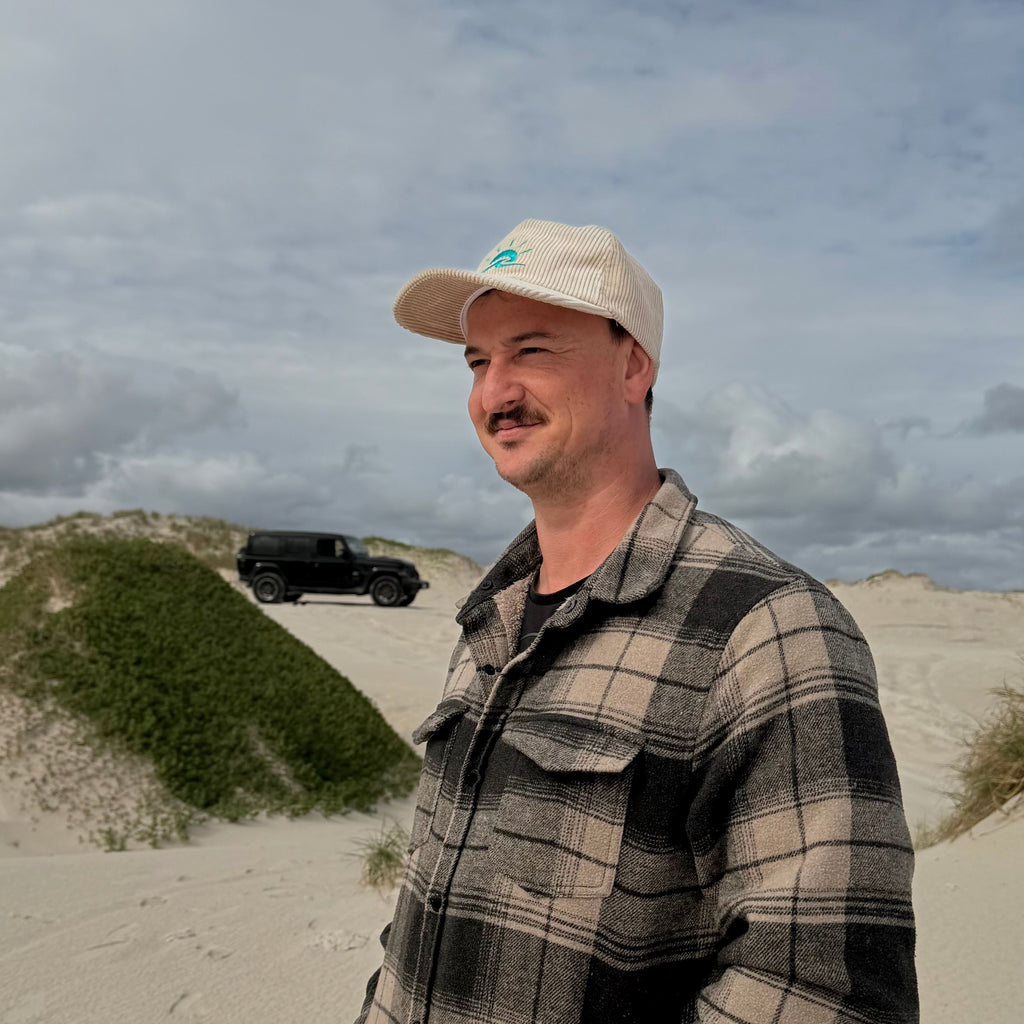 Man standing at beach, 4WD in background, wearing Aquaholics corduroy hat